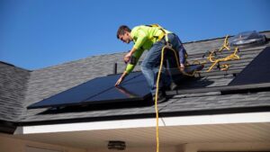 A person inspects a sunlit residential roof with trees and power lines in the background.