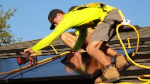 A worker in safety harness installing solar panels on a residential shingle roof under bright daylight.