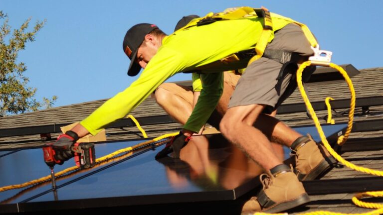 A worker in safety harness installing solar panels on a residential shingle roof under bright daylight.